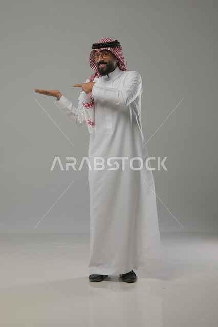 ارتداء الثوب والشماغ السعودي التقليدي  , بورتريه لرجل عربي خليجي سعودي مبتسم يفتح كفه للأعلى ويشير إلى عنصر ما ،النظر إلى الكاميرا  ،صورة الجسم بالطول الكامل ، خلفية رمادية