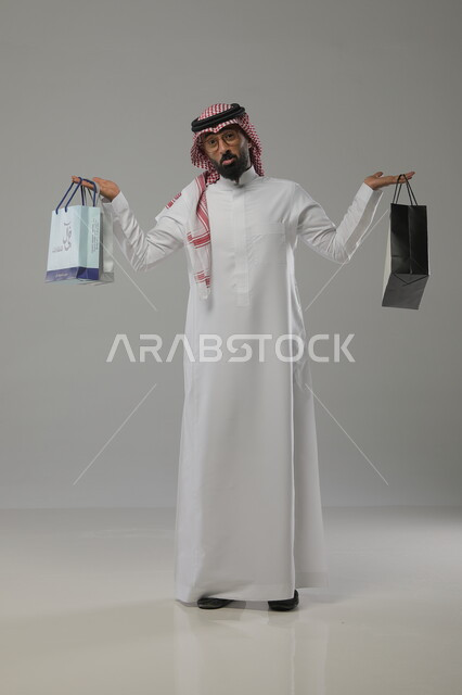الاستفادة من العروض والخصومات الموسمية  ,  قضاء أوقات ممتعة في التبضع والشراء  , بورتريه لرجل عربي سعودي خليجي مبتسم يرتدي الثوب والشماغ التقليدي يرفع يديه نحو الأعلى ويحمل بيده أكياس التسوق , صورة الجسم بالطول الكامل, خلفية رمادية
