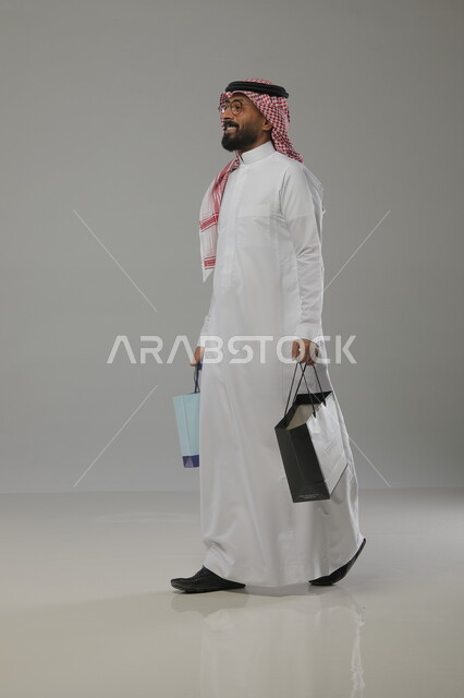 العودة من المول التجاري , قضاء أوقات ممتعة في الشراء و التبضع , الاستفادة من العروض والخصومات  الموسمية , بورتريه لرجل عربي سعودي خليجي يرتدي الثوب والشماغ  التقليدي ويحمل بيده أكياس التسوق , صورة الجسم بالطول الكامل, خلفية رمادية
