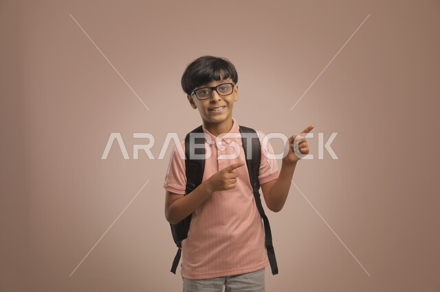 الإشارة بإصبعيه إلى شيء ما  ، الوقوف باستقامة والنظر إلى الكاميرا  , بورتريه لطالب عربي خليجي سعودي مبتسم  يرتدي الزي الكاجوال و يحمل الحقيبة المدرسية، الإستعداد للعام  الجديد، الشعور بالحماس والنشاط في بداية السنة الدراسية ، خلفية بنية