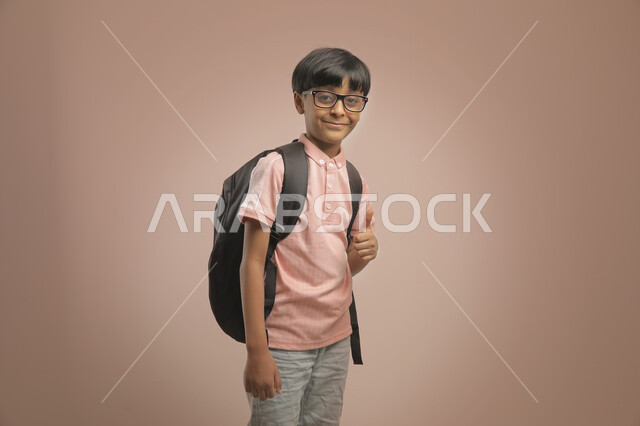 رفع الإبهام لاعلى للشعور بالرضى والاعجاب بالعودة إلى المدارس ، الوقوف باستقامة والنظر إلى الكاميرا  , بورتريه لطالب عربي خليجي سعودي يرتدي الزي الكاجوال و يحمل الحقيبة المدرسية، الشعور بالحماس والنشاط ببداية العام الدراسي  ، خلفية بنية