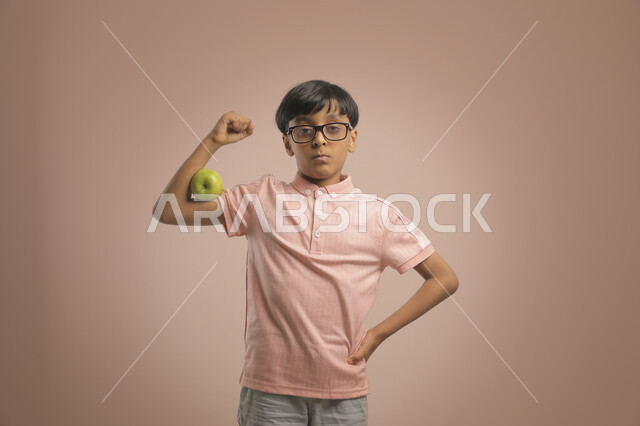 التمتع ببنية جسدية قوية ,  إيماءات القوة والصحة والرشاقة  , بورتريه لولد عربي خليجي سعودي يرتدي النضارات الطبية و الزي الكاجوال  ينظر إلى الكاميرا ،  تناول فاكهة التفاح المغذي  ، خلفية بنية