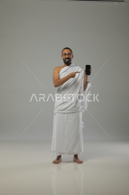 أداء العبادات الطاعات والتقرب الى الله، الدليل الالكتروني للحاج والمعتمر, عرض تطبيقات الهاتف المحمول لارشادات الحج و العمرة, بورتريه لرجل عربي خليجي سعودي يرتدي لباس الاحرام والنظارات الطبية يحمل بيده الجوال، صورة الجسم بالطول الكامل، خلفية رمادية