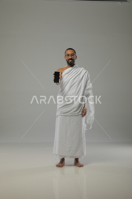الدليل الالكتروني للحاج والمعتمر, عرض تطبيقات الهاتف المحمول لارشادات الحج و العمرة, بورتريه لرجل عربي خليجي سعودي يرتدي لباس الاحرام والنظارات الطبية يحمل بيده الجوال، أداء العبادات الطاعات والتقرب الى الله، صورة الجسم بالطول الكامل، خلفية رمادية