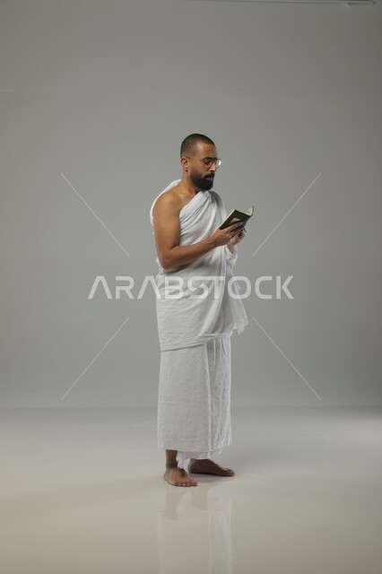 آداء فريضة الحج، العبادة والدعاء والتقرب الى الله، بورتريه لرجل مسلم عربي سعودي خليجي يرتدي ملابس الاحرام والنظارات الطبية ويحمل بيده القرآن الكريم، موسم العمرة في شهر رمضان المبارك،صورة الجسم بالطول الكامل ، خلفية رمادية