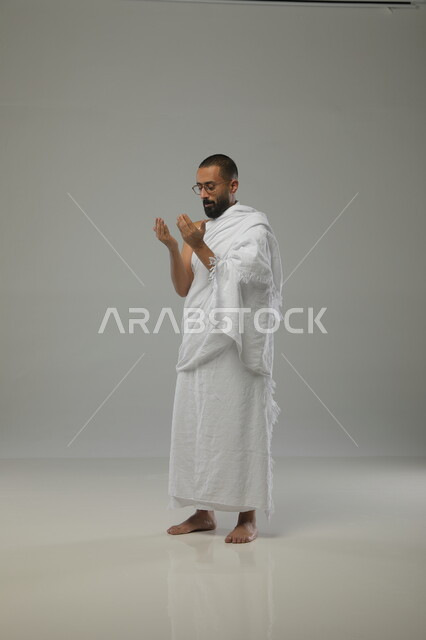 Supplication, worship, and getting closer to God, a side portrait of a Saudi Arabian Gulf Muslim man wearing Ihram clothing and medical glasses, praying with supplication and reverence and raising his hands up, performing the Hajj pilgrimage, the Umrah season in the holy month of Ramadan, full-length body portrait, gray background