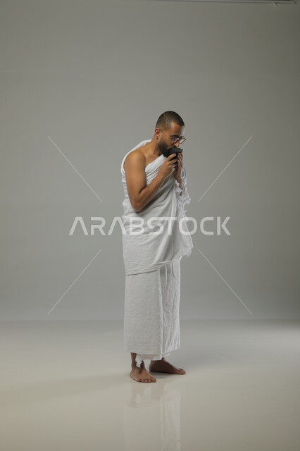 قراءة القرآن الكريم ، الدعاء والعباده والتقرب الى الله، بورتريه جانبي لرجل مسلم عربي سعودي خليجي يرتدي ملابس الاحرام ويقوم بآداء فريضة الحج، موسم العمرة في شهر رمضان المبارك، صورة الجسم بالطول الكامل ،  خلفية رمادية