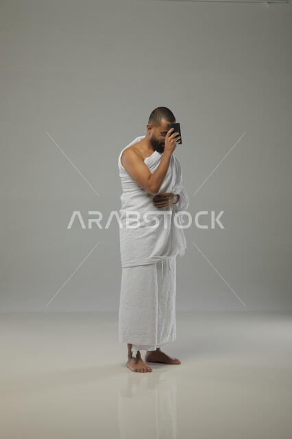 الدعاء والعباده والتقرب الى الله، بورتريه جانبي لرجل مسلم عربي سعودي خليجي يرتدي ملابس الاحرام ويقوم بآداء فريضة الحج، موسم العمرة في شهر رمضان المبارك، قراءة القرآن الكريم  ، صورة الجسم بالطول الكامل ،خلفية رمادية