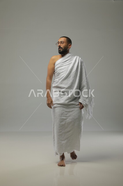 التقرب الى الله بالدعاء والعبادة ، بورتريه جانبي لرجل مسلم عربي سعودي خليجي يرتدي ملابس الاحرام وآداء فريضة الحج، موسم العمرة في شهر رمضان المبارك، صورة الجسم بالطول الكامل ،خلفية رمادية