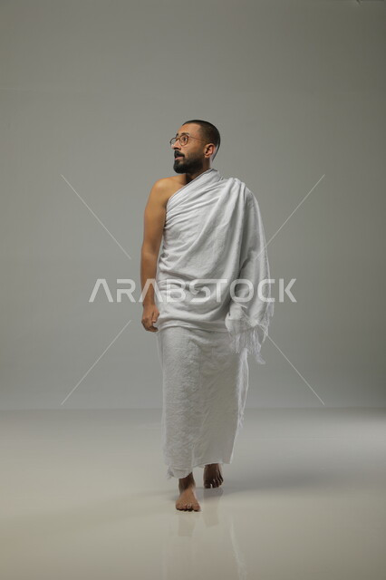 العباده والدعاء والتقرب الى الله، بورتريه جانبي لرجل مسلم عربي سعودي خليجي يرتدي ملابس الاحرام ويقوم بآداء فريضة الحج، موسم العمرة في شهر رمضان المبارك،صورة الجسم بالطول الكامل ، خلفية رمادية