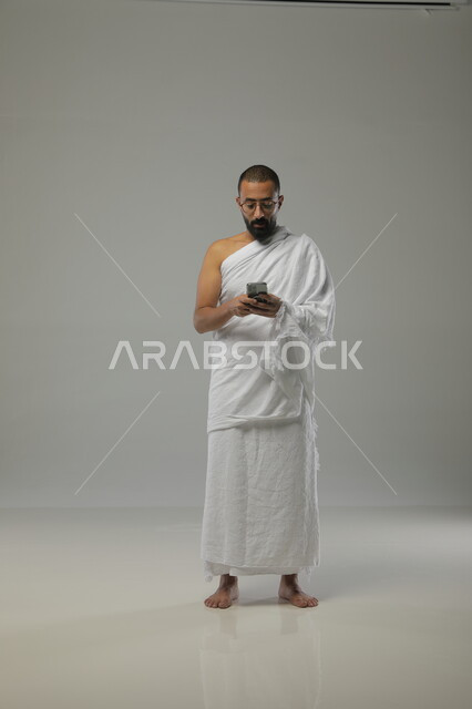 دليل المعتمر الالكتروني، استخدام تطبيق ارشادات مناسك الحج والعمرة، بورتريه لرجل عربي خليجي سعودي يرتدي ملابس الاحرام والنظارات الطبية يقوم باستخدام الهاتف المحمول، العبادات والتقرب الى الله، قراْة القرآن الكريم عن طريق الجوال، خلفية رمادية