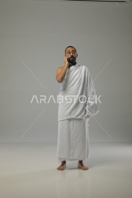 إجراء مكالمة هاتفية، بورتريه لرجل عربي خليجي سعودي يرتدي ملابس الاحرام ونظارات طبية يقوم بالرد على اتصالات الهاتف المحمول، اداء مناسك الحج والعمرة، طاعات  وعبادات التقرب الى الله، صورة الجسم بالطول الكامل، خلفية رمادية