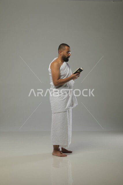 آداء مناسك الحج والعمرة، تلاوة الآيات والتقرب الى الله، حفظ وتفسير القرآن الكريم، بورتريه جانبي لرجل مسلم عربي خليجي سعودي يرتدي ملابس الاحرام ونظارات طبية يحمل بيده مصحف صغير، الركن الخامس من اركان السلام،  صورة الجسم بالطول الكامل، خلفية رمادية
