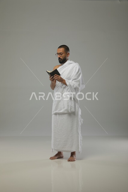 تلاوة الآيات والتقرب الى الله، حفظ وتفسير القرآن الكريم، بورتريه لرجل مسلم عربي خليجي سعودي يرتدي ملابس الاحرام ونظارات طبية يحمل بيده مصحف صغير، آداء مناسك الحج والعمرة، الركن الخامس من اركان السلام،  صورة الجسم بالطول الكامل، خلفية رمادية