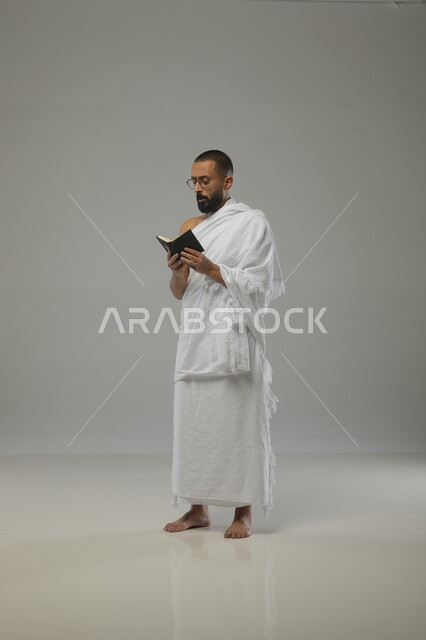 تلاوة الآيات والتقرب الى الله، حفظ وتفسير القرآن الكريم، بورتريه لرجل مسلم عربي خليجي سعودي يرتدي ملابس الاحرام ونظارات طبية يحمل بيده مصحف صغير، آداء مناسك الحج والعمرة، الركن الخامس من اركان السلام،  صورة الجسم بالطول الكامل، خلفية رمادية