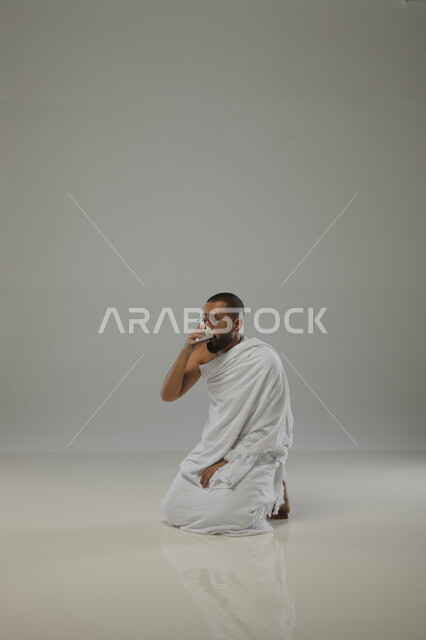 Drinking water while sitting in three batches, drinking and quenching yourself from Zamzam water, supplication, worship, and getting closer to God, a side portrait of a Saudi Arabian Gulf Muslim man wearing Ihram clothing, following the Sunnah of the Messenger, performing Hajj and Umrah rituals, gray background.