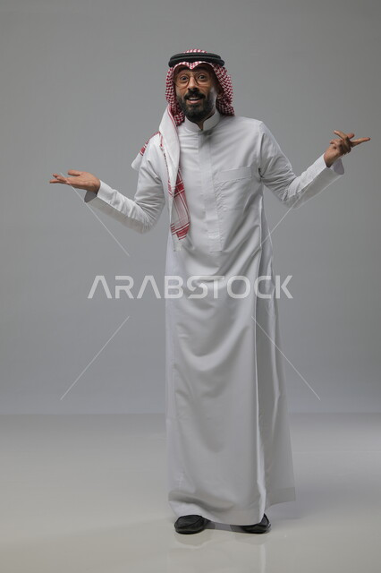 التساؤل والاستفهام والاستفسار عن شيء ما، ايماءات عدم المعرفة والدهشة والاستغراب، بورتريه لرجل عربي خليجي سعودي يرتدي الشماغ والثوب التقليدي والنظارات الطبية يرفع يديه للاعلى، صورة الجسم بالطول الكامل، خلفية رمادية
