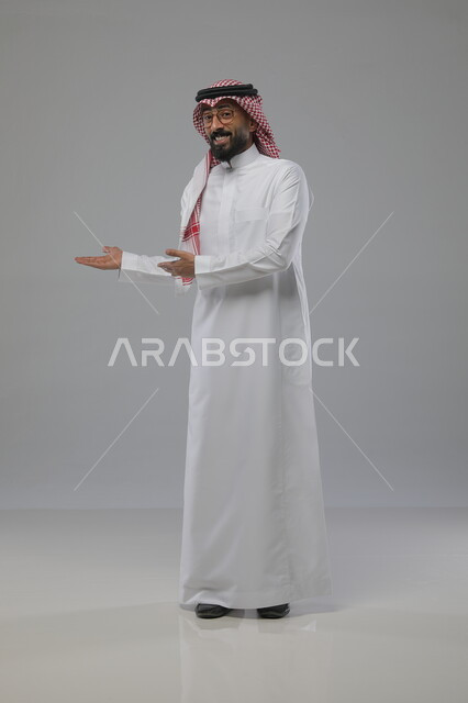 حركات وايماءات تدل على هذا المكان وهذا الموقع, الاشارة الى مكان ما، بورتريه لرجل عربي خليجي سعودي مبتسم يرتدي الشماغ والثوب التقليدي والنظارات الطبية ينظر الى الكاميرا، الترحيب بشخص ما، صورة الجسم بالطول الكامل، خلفية رمادية