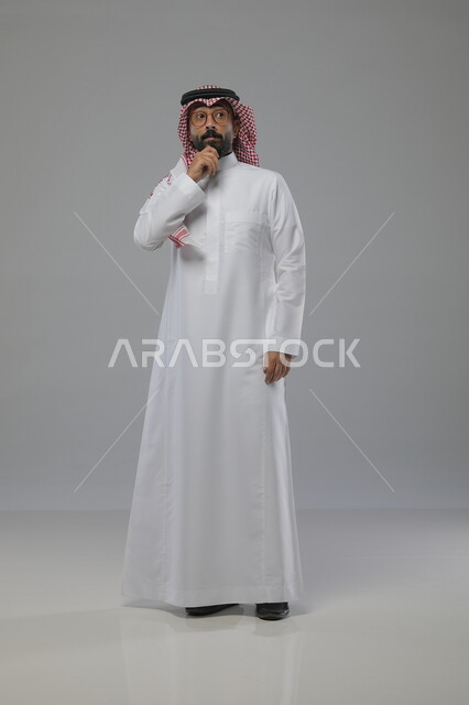 تعابير عدم المعرفة والدهشة والاستغراب، التساؤل والاستفهام والاستفسار عن شيء ما، بورتريه لرجل عربي خليجي سعودي يرتدي الشماغ والثوب التقليدي والنظارات الطبية يضع يده على ذقنه بايماءات التفكير، صورة الجسم بالطول الكامل، خلفية رمادية