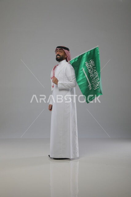 يوم العلم 11 مارس، ذكرى اليوم الوطني السعودي 23 سبتمبر، بورتريه لرجل عربي خليجي سعودي يرتدي الشماغ والثوب التقليدي ونظارات طبية يحمل بيده علم السعودية، الاحتفال بالعيد الوطني، الفخر والاعتزاز بامتلاك الهوية الوطنية، صورة الجسم بالطول الكامل، خلفية رمادية