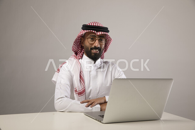 إجراء مقابلات عمل أونلاين, قضاء أوقات ممتعه بتصفح وسائل التواصل الاجتماعي باستخدام الحاسب المحمول, انجاز الأعمال عن بعد, بورتريه لرجل عربي خليجي سعودي مبتسم يرتدي الشماغ والثوب التقليدي والنظارات الطبية ينظر إلى الكاميرا بإيماءات السرور والسعاده, خلفيه رماديه