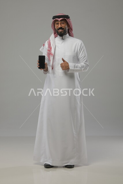 مفهوم التسويق الرقمي الالكتروني، عرض تطبيقات وخدمات الهاتف المحمول، بورتريه لرجل عربي خليجي سعودي مبتسم يرتدي االشماغ ولثوب التقليدي والنظارات الطبية يحمل بيده جوال بشاشة سوداء فارغة يرفع اصبع الابهام للدلالة على الاعجاب، صورة الجسم بالطول الكامل، خلفية رمادية