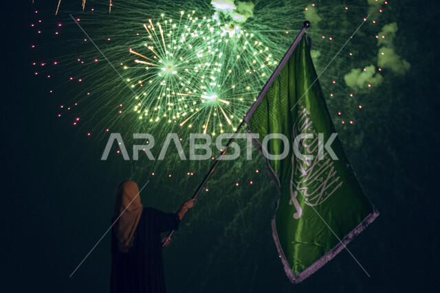 صورة من الخلف لمواطنة عربية خليجية سعودية ترفع علم المملكة في احتفالات اليوم الوطني 23 سبتمبر، الالعاب النارية الخضراء في سماء المملكة احتفالا بذكرى يوم العلم 11 مارس، العيد الوطني السعودي، الفخر والاعتزاز بالانتماء للسعودية