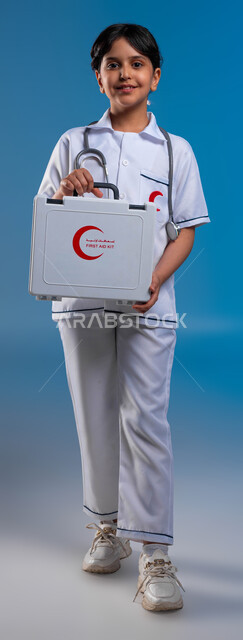 النظر إلى الكاميرا، آمال مستقبلية في الوصول للحلم، بورتريه لفتاة عربية خليجية سعودية مبتسمة ترتدي زي الطبيب وتحمل بيدها حقيبة الاسعافات الأولية تحلم أن تصبح طبيبة محترفه، مساعدة المرضى وطريقة التعامل في الحالات الطارئة، صورة الجسم بالطول الكامل، خلفية زرقاء