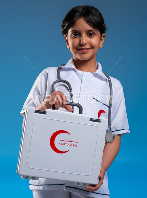 أمنيات و آمال مستقبلية في الوصول للحلم ،النظر إلى الكاميرا، التعامل مع حالات الطوارئ، بورتريه مقرب لفتاة عربية خليجية سعودية مبتسمة ترتدي زي الطبيب وتحمل بيدها حقيبة الاسعافات الأولية تحلم أن تصبح طبيبة محترفه، مساعدة المرضى في الحالات الطارئة، خلفية زرقاء