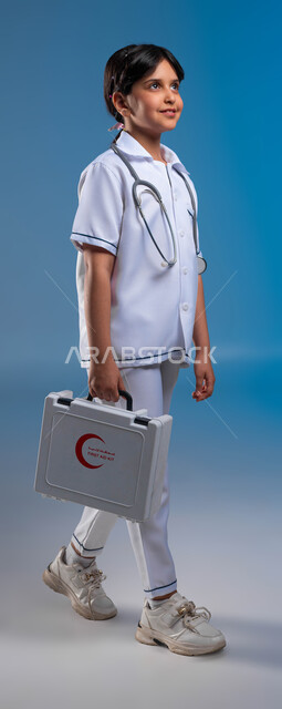 مساعدة المرضى في الحالات الطارئة، الطموحات و الأهداف المستقبلية في العمل في المستشفيات ،بورتريه مقرب لفتاة عربية خليجية سعودية مبتسمة ترتدي زي الطبيب تحمل بيدها حقيبة الاسعافات الاولية تحلم أن تصبح طبيبة محترفه, صورة الجسم بالطول الكامل، خلفية زرقاء