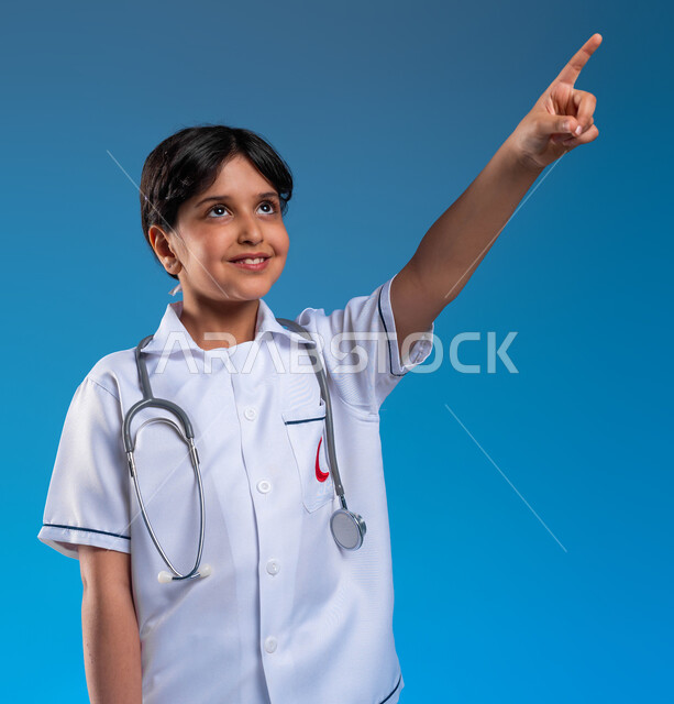 Future ambitions and goals for working in hospitals, a close-up portrait of a smiling Saudi Gulf Arab girl wearing a doctor’s uniform, dreaming of becoming an ambulance and emergency doctor, the concept of health care, defining the profession since childhood, blue background