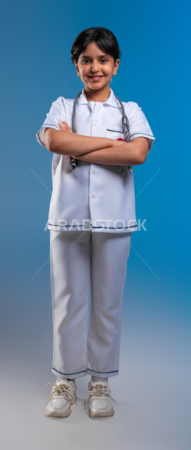 الأهداف والطموحات المستقبلية في العمل في المستشفيات ،الوقوف بإيماءات أيدي متقاطعه و النظر إلى الكاميرا، بورتريه لفتاة عربية خليجية سعودية مبتسمة ترتدي زي الطبيب تحلم أن تصبح طبيبة إسعاف وطوارئ, تحديد المهنة منذ الصغر، صورة الجسم بالطول الكامل، خلفية زرقاء