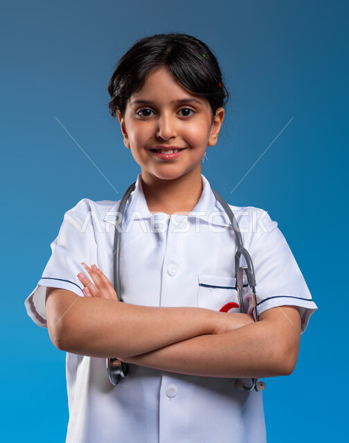 الوقوف بإيماءات أيدي متقاطعه و النظر إلى الكاميرا، الطموحات و الأهداف المستقبلية في العمل في المستشفيات ،بورتريه مقرب لفتاة عربية خليجية سعودية مبتسمة ترتدي زي الطبيب تحلم أن تصبح طبيبة إسعاف وطوارئ, مفهوم الرعاية الصحية، تحديد المهنة منذ الصغر، خلفية زرقاء