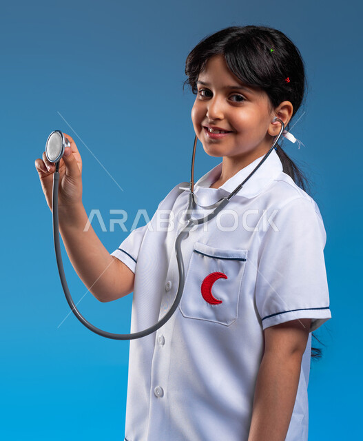 النظر إلى الكاميرا بإيماءات الشغف بتحقيق حلم الطفولة، استخدام سماعة نبضات القلب، بورتريه مقرب لفتاة عربية خليجية سعودية مبتسمة ترتدي زي الطبيب تحلم أن تصبح طبيبة محترفة، اختيار وتحديد المهنة المستقبلية منذ الصغر، طموحات وأهداف طفولية، خلفية زرقاء