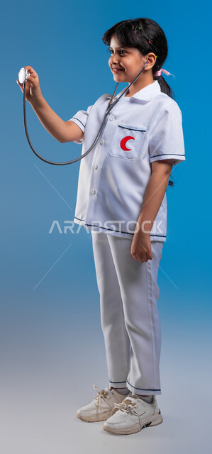طموحات وأهداف طفولية، استخدام سماعة نبضات القلب، ايماءات الشغف بتحقيق حلم الطفولة، بورتريه لفتاة عربية خليجية سعودية مبتسمة ترتدي زي الطبيب تحلم أن تصبح طبيبة محترفة، اختيار وتحديد المهنة المستقبلية منذ الصغر، صورة الجسم بالطول الكامل، خلفية زرقاء
