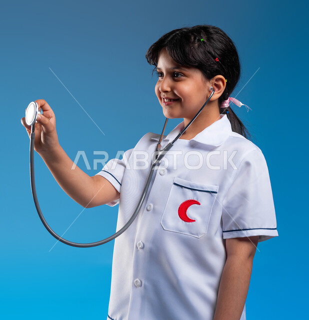 Using a heartbeat stethoscope, gestures of passion for achieving a childhood dream, a close-up portrait of a smiling Saudi Arabian Gulf girl wearing a doctor’s uniform dreaming of becoming a professional doctor, the concept of medicine and health care, choosing and defining a future profession since childhood, childhood ambitions and goals, blue background
