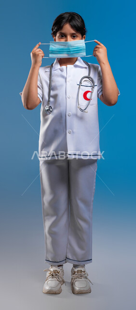 طموحات مستقبلية في العمل في المستشفيات، الاحترازات الوقائية ضد كوفيد 19 ،مفهوم الطب والرعاية الصحية، بورتريه مقرب لفتاة عربية خليجية سعودية ترتدي زي الطبيب و تقوم بوضع الكمامة ، تحديد مهنة المستقبل منذ الصغر، صورة الجسم بالطول الكامل، خلفية زرقاء