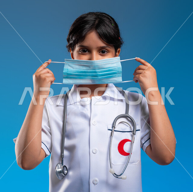 الاحترازات الوقائية ضد كوفيد 19 ،مفهوم الطب والرعاية الصحية، طموحات وأهداف مستقبلية في العمل في المستشفيات، بورتريه مقرب لفتاة عربية خليجية سعودية ترتدي زي الطبيب و تقوم بوضع الكمامة ، اختيار وتحديد مهنة المستقبل منذ الصغر، خلفية زرقاء