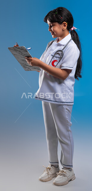 إعداد التقارير الطبيه، تحديد مهنة المستقبل منذ الصغر، الأهداف المستقبلية في العمل في العيادات والمشافي ،بورتريه لفتاة عربية خليجية سعودية مبتسمة ترتدي اللباس الابيض الخاص بمهنة الطب و تقوم بتدوين نتائج الفحوصات بالسجل الطبي، صورة الجسم بالطول الكامل، خلفية زرقاء