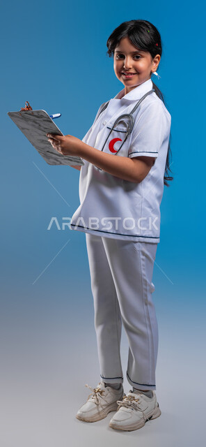 تحديد مهنة المستقبل منذ الصغر، تدوين نتائج الفحوصات بالسجل الطبي، الأهداف المستقبلية في العمل في العيادات والمشافي ،بورتريه لفتاة عربية خليجية سعودية مبتسمة ترتدي اللباس الابيض الخاص بمهنة الطب و تقوم بإعداد التقارير الطبيه، صورة الجسم بالطول الكامل، خلفية زرقاء
