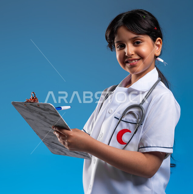 تدوين نتائج الفحوصات بالسجل الطبي، الطموحات و الأهداف المستقبلية في العمل في العيادات والمشافي ،بورتريه مقرب لفتاة عربية خليجية سعودية مبتسمة ترتدي اللباس الابيض الخاص بمهنة الطب و تقوم بإعداد التقارير الطبيه، تحديد واختيار مهنة المستقبل منذ الصغر، خلفية زرقاء