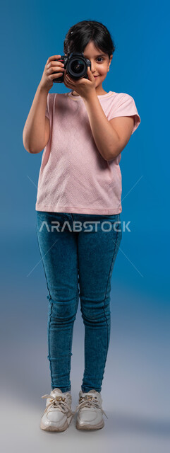 الوقوف بثقة والتقاط صور تذكارية, بورتريه لفتاة عربية خليجية سعودية ترتدي زي الكاجوال تمسك كاميرا التصوير الفوتوغرافي, توثيق اللحظات السعيدة والاستمتاع بممارسة مهنة المصورة المحترفة, الأحلام والأمنيات المستقبلية للبنات, صورة الجسم بالطول الكامل, خلفية زرقاء
