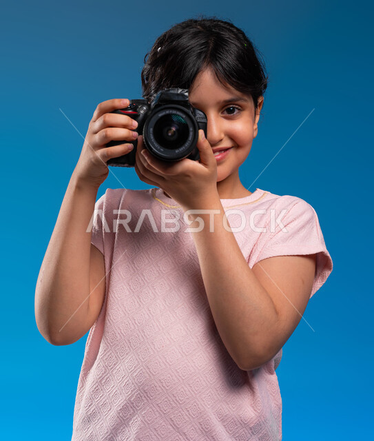 الأحلام والأمنيات المستقبلية للبنات, الوقوف بثقة والتقاط صور تذكارية, بورتريه مقرب لفتاة عربية خليجية سعودية ترتدي زي الكاجوال تمسك كاميرا التصوير الفوتوغرافي, توثيق اللحظات السعيدة والاستمتاع بممارسة مهنة المصورة المحترفة, خلفية زرقاء