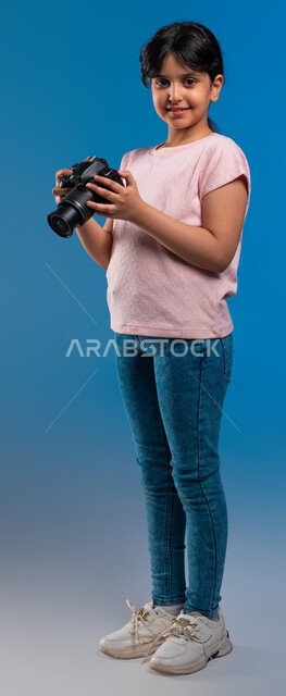 توثيق اللحظات السعيدة وممارسة هواية التصوير, التقاط الصور التذكارية, بورتريه لفتاة عربية خليجية سعودية ترتدي زي الكاجوال تمسك الكاميرا الفوتوغرافية, تحقيق الأحلام المستقبلية للحصول على مهنة مصورة محترفة, صورة الجسم بالطول الكامل, خلفية زرقاء