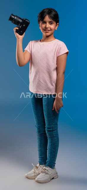 الشغف لتوثيق اللحظات السعيدة والاستمتاع بممارسة مهنة المصورة المحترفة, الوقوف بثقة والتقاط صور تذكارية, بورتريه لفتاة عربية خليجية سعودية ترتدي زي الكاجوال تمسك كاميرا التصوير الفوتوغرافي, الأحلام المستقبلية للبنات, صورة الجسم بالطول الكامل, خلفية زرقاء