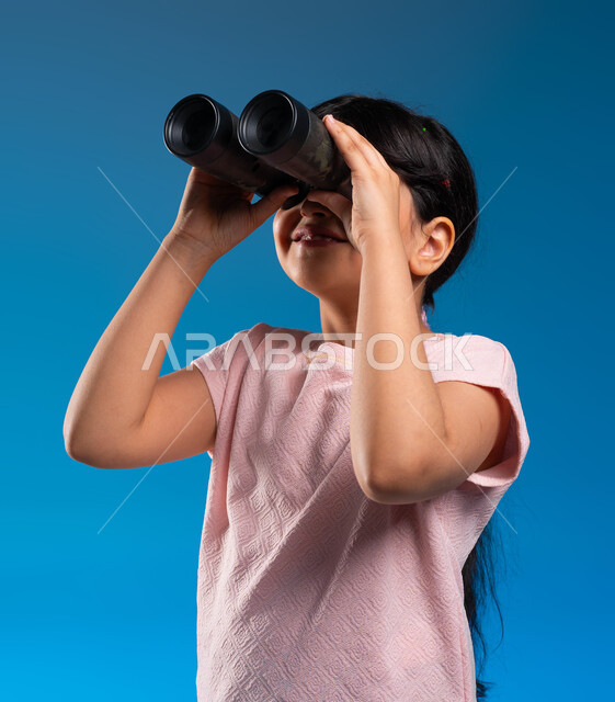 التخطيط المستقبلي للأهداف والطموحات، حب الاستكشاف والمغامرة، التركيز على شيء ما، رؤية الاشياء البعيدة، بورتريه مقرب لفتاة عربية خليجية سعودية تمسك بيديها منظار الرؤية ثنائي العينين لمشاهدة شيئا ما، خلفية زرقاء