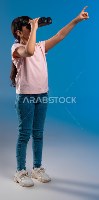 رؤية الاشياء البعيدة، أهداف وطموحات مستقبلية، بورتريه  لفتاة عربية خليجية سعودية تمسك بيدها منظار الرؤية ثنائي العينين لمشاهدة شيئا ما وتشير بيدها الأخرى تجاه الهدف، متعة الاستكشاف والمغامرة، صورة الجسم بالطول الكامل، خلفية زرقاء
