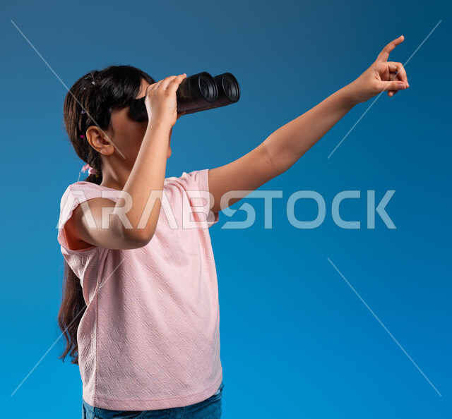 أهداف وطموحات مستقبلية، رؤية الاشياء البعيدة، بورتريه مقرب لفتاة عربية خليجية سعودية تمسك بيدها منظار الرؤية ثنائي العينين وتشير بيدها الأخرى تجاه هدف ما، متعة الاستكشاف والمغامرة، خلفية زرقاء