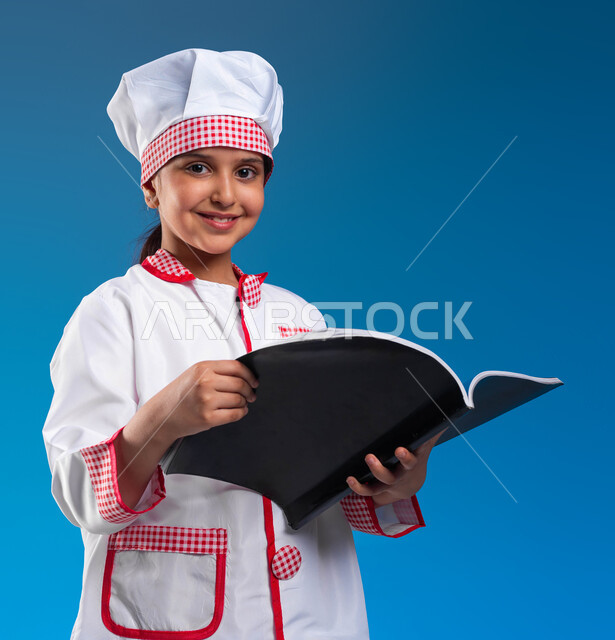 احتراف مهنة الطهي، بورتريه مقرب لفتاة عربية خليجية سعودية ترتدي زي الطباخه تحمل بيدها كتاب وتنظر الى الكاميرا بابتسامه، تطبيق وصفات الطعام من كتب الطبخ، طموحات وأهداف مستقبيلة، فنون الأكل و مهارات الإبتكار والتذوق، خلفية زرقاء