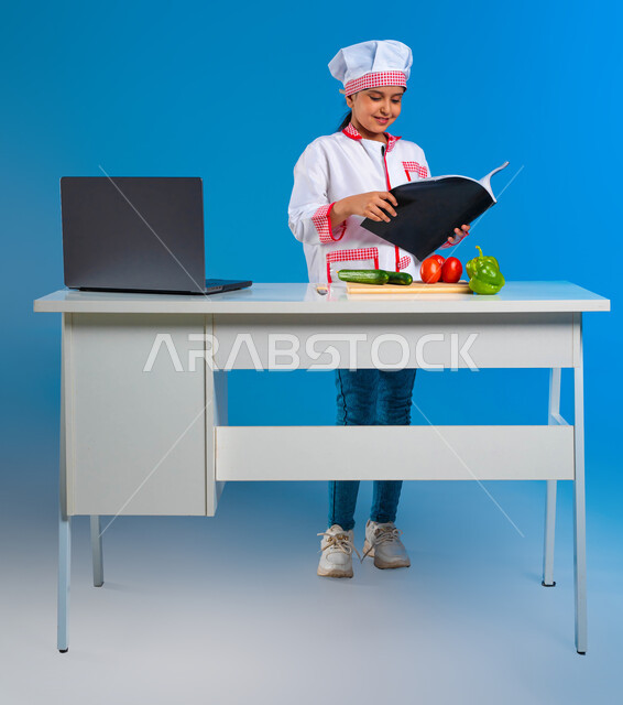 الابتكار لتحقيق الأحلام المستقبلية, البشاشة والشغف وحب تعلم فنون الطبخ, بورتريه لفتاة عربية خليجية سعودية ترتدي زي الشيف تمسك كتاب إعداد الطعام بإيماءات السرور, تعلم وصفات جديده صحية, احتراف مهنة الطهي, خلفية زرقاء